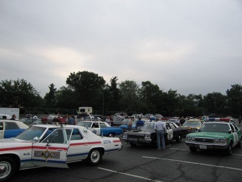 1991 Ford LTD Crown Victoria - lot of vintage police cars at dusk, including a white State Police cruiser marked 1776 and a green-and-white sedan, with onlookers.