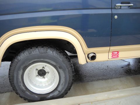 1986 Ford Bronco - Close-up side view of a blue and tan SUV, showing the rear wheel, exhaust pipe, and a "Fire Extinguisher Inside" sticker.
