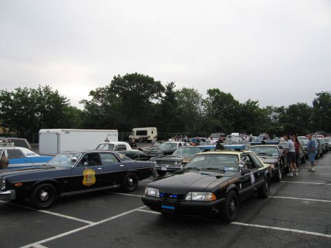 1991 Ford LTD Crown Victoria - black police Mustang and a black police sedan with a gold door shield parked among vintage cruisers as people walk between rows.