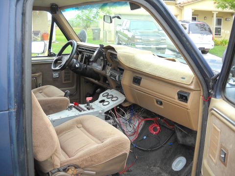 1986 Ford Bronco - The interior of a blue truck shows tan seats and a custom center console with exposed wiring, with other vehicles visible through the winds…
