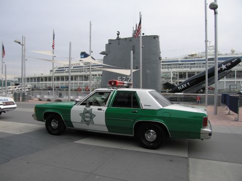 1991 Ford LTD Crown Victoria - green-and-white sheriff sedan with a star shield parked on a waterfront plaza in front of a museum submarine and Navy display.