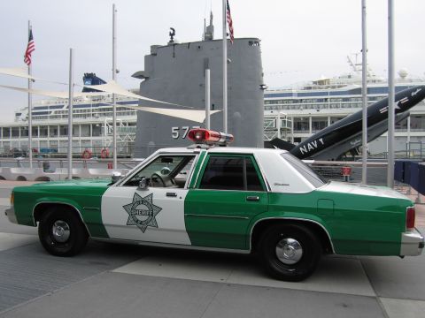 1991 Ford LTD Crown Victoria - Side profile of a green-and-white sheriff sedan with a single red roof light parked beside a museum submarine numbered 57.