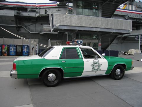 1991 Ford LTD Crown Victoria - Side view of a green-and-white sheriff sedan with a roof beacon parked on a plaza beside blue vending machines and a hangar deck.