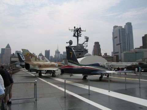 1991 Ford LTD Crown Victoria - camouflage fighter jet and a blue-and-white jet displayed on an aircraft carrier flight deck with a city skyline in the background.