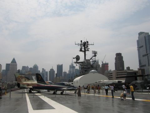 1991 Ford LTD Crown Victoria - Vintage military jets and a helicopter parked on an aircraft carrier flight deck, with the carrier island superstructure and Manhattan skyl…