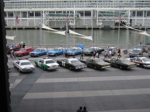 1991 Ford LTD Crown Victoria - long row of classic American police cruisers in various state and local liveries parked along a waterfront pier in front of a glass termina…