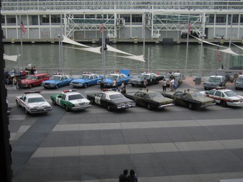 1991 Ford LTD Crown Victoria - Classic American police cruisers and a small blue convertible arranged in a row on a waterfront pier with a glass building behind.