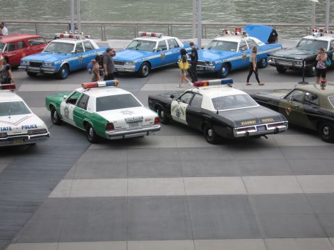 1991 Ford LTD Crown Victoria - Several restored vintage police cars on a paved plaza, including a green-and-white Sheriff car and a black-and-white Highway Patrol cruiser.
