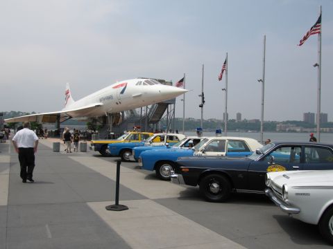 1991 Ford LTD Crown Victoria - British Airways Concorde on display behind a row of classic blue and tan police cars lined up along a waterfront pier under U.S. flags.