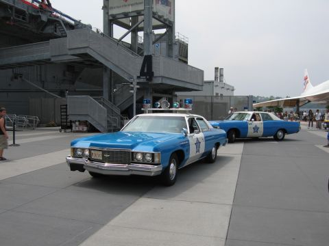 1991 Ford LTD Crown Victoria - Two classic blue-and-white police sedans with star-shaped door badges parked on a paved plaza beside a museum building, Concorde in the dis…