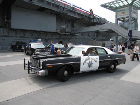 1991 Ford LTD Crown Victoria - black-and-white Highway Patrol sedan with a chrome push bar and roof lights parked on a plaza, with other police cars and a carrier hull be…