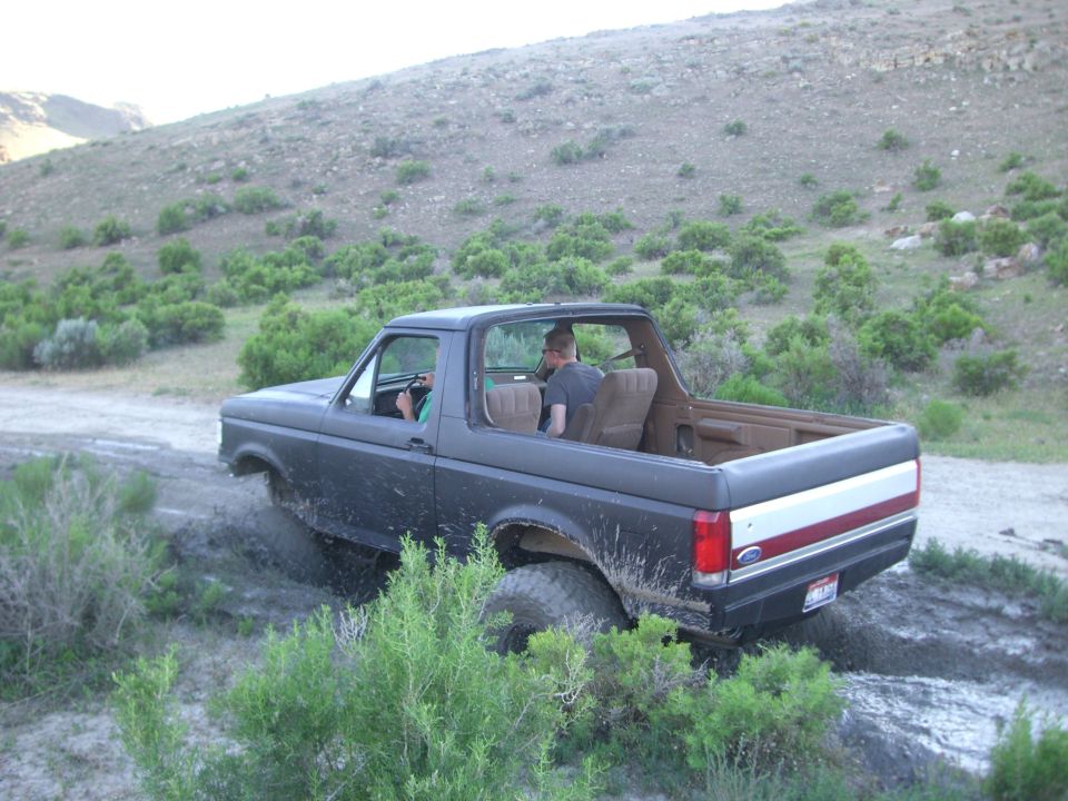 Dark Ford Bronco driving through mud and brush on a dirt road with a hill in the background
