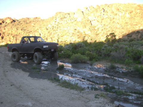 1987 Ford Bronco - A modified black truck navigates a muddy trail in a dry, hilly landscape.