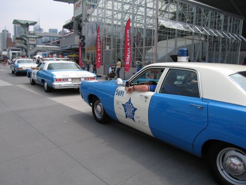 1991 Ford LTD Crown Victoria - receding line of vintage blue-and-white police cars parked along a pier next to a glass museum building, the nearest marked with number 569…