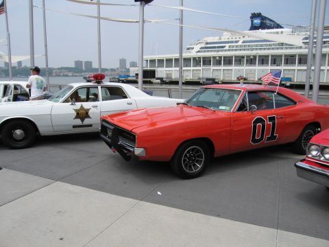 1991 Ford LTD Crown Victoria - orange muscle car bearing a large 01 and a small flag parked beside a white Sheriff cruiser on a waterfront pier, cruise ship in the backgr…