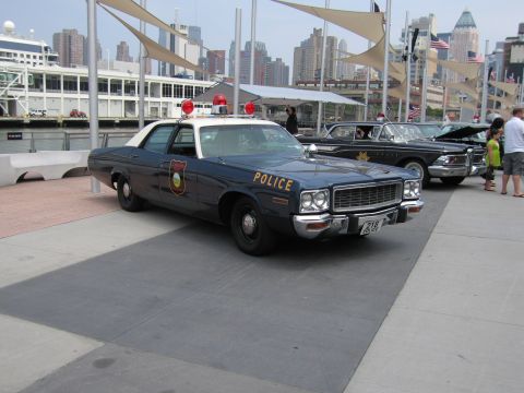 1991 Ford LTD Crown Victoria - dark blue-and-white vintage Police sedan with red roof lights parked on a paved waterfront plaza, with the NYC skyline and other cruisers b…