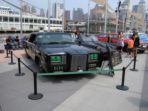 1991 Ford LTD Crown Victoria - glossy black custom car with distinctive green-glowing headlights displayed behind a roped barrier on a waterfront plaza, NYC skyline behin…