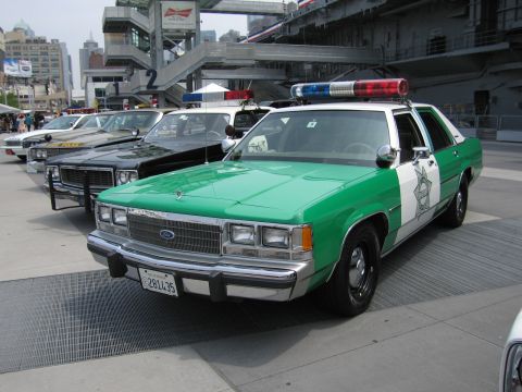 1991 Ford LTD Crown Victoria - green-and-white Ford police sedan with roof light bar at the front of a row of vintage cruisers parked beneath an aircraft carrier's overha…