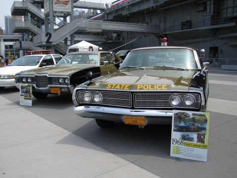 1991 Ford LTD Crown Victoria - Two olive-and-black State Police sedans displayed with a placard reading 1968 Ford Custom 500 on a plaza beneath an aircraft carrier overha…