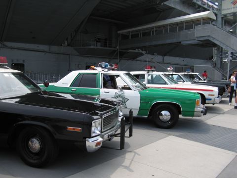1991 Ford LTD Crown Victoria - black police cruiser with push bar beside green-and-white and black-and-white Sheriff cars parked in a row beneath an aircraft carrier stru…