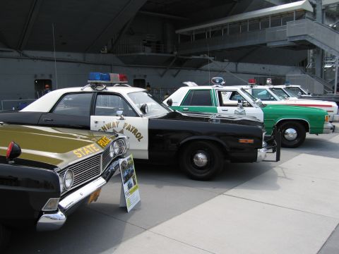 1991 Ford LTD Crown Victoria - row of vintage police cars including an olive State Police sedan, a black-and-white Highway Patrol car and green-and-white cruisers under a…