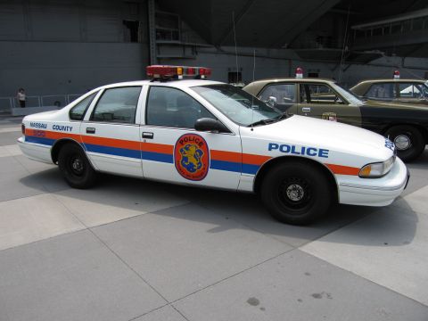 1991 Ford LTD Crown Victoria - white Nassau County Police sedan with orange and blue stripes and a circular department seal, roof light bar, parked under a carrier overha…