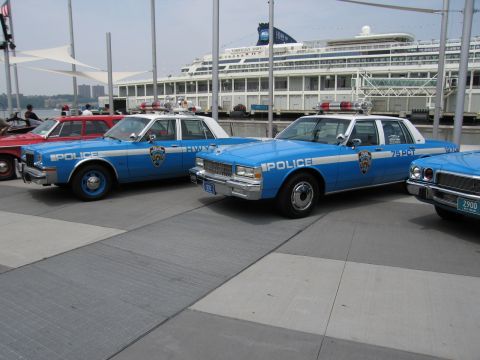 1991 Ford LTD Crown Victoria - row of vintage blue-and-white NYPD Police sedans with roof light bars parked along a pier, cruise ship and red car at the edge of frame.