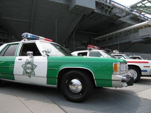 1991 Ford LTD Crown Victoria - Front three-quarter view of a green-and-white Sheriff sedan with a star door emblem and roof light bar, other cruisers behind under a carri…