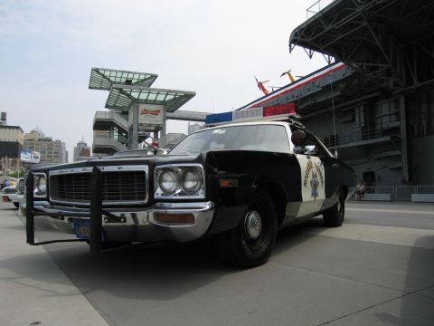 1991 Ford LTD Crown Victoria - Front three-quarter view of a black and white Dodge highway patrol cruiser with push bar and light bar on an outdoor plaza near buildings.