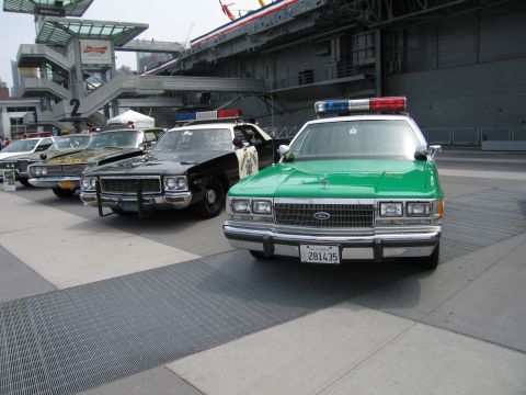 1991 Ford LTD Crown Victoria - green Ford police sedan leads a row of vintage cruisers including a black and white car, displayed outdoors at a public show.