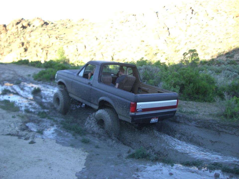 1987 Ford Bronco driving through a muddy trail with two people inside
