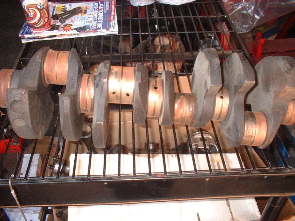 Engine crankshaft with copper journals lying on a black wire shelf in a shop