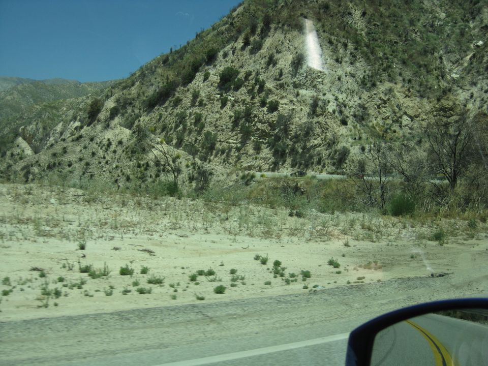 Desert landscape with a road winding through hills under a clear blue sky. A car's side mirror is visible in the foreground