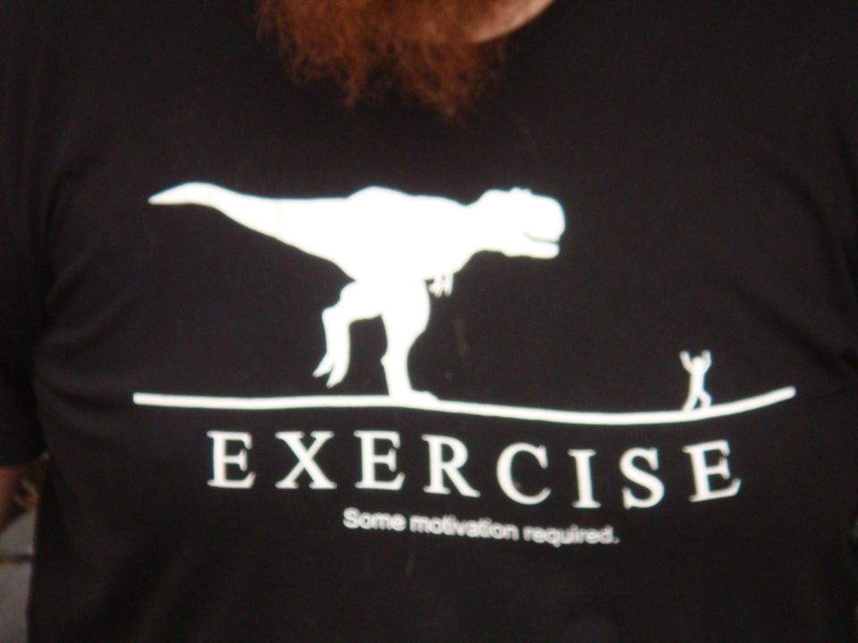 Black t-shirt with white graphic of a T-Rex and a stick figure on a line, text reads EXERCISE Some motivation required