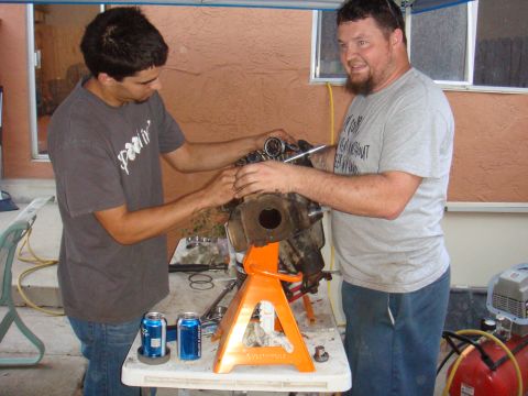 1992 Ford Bronco - Two men work on an engine part supported by jack stands, with tools and drinks nearby.