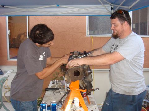 1992 Ford Bronco - Two men are seen working on a vehicle axle assembly that is supported by an orange jack stand.