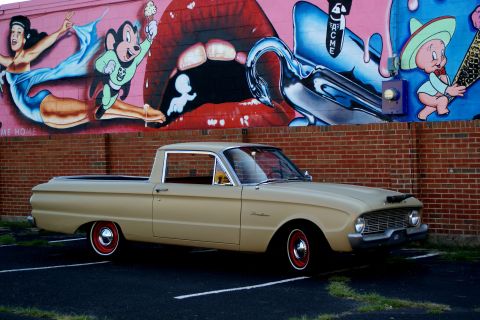 1972 Ford Ranchero - Side profile of a tan early-1960s Ford Falcon Ranchero car-based pickup parked on asphalt in front of a brick wall topped by a colorful car…