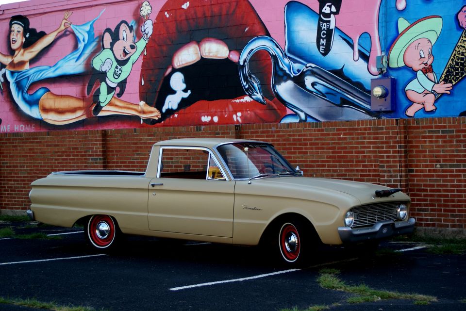 Tan early-60s Ford Ranchero in profile parked before a brick wall with a cartoon mural