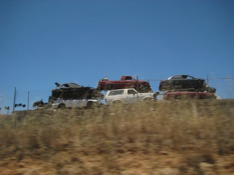 1996 Ford Bronco - A white 1996 Ford Bronco is visible among several other vehicles stacked in a junkyard under a clear blue sky.