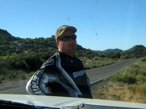 1996 Ford Bronco - A man in a racing jersey stands by a road in a dry, hilly landscape under a clear blue sky.