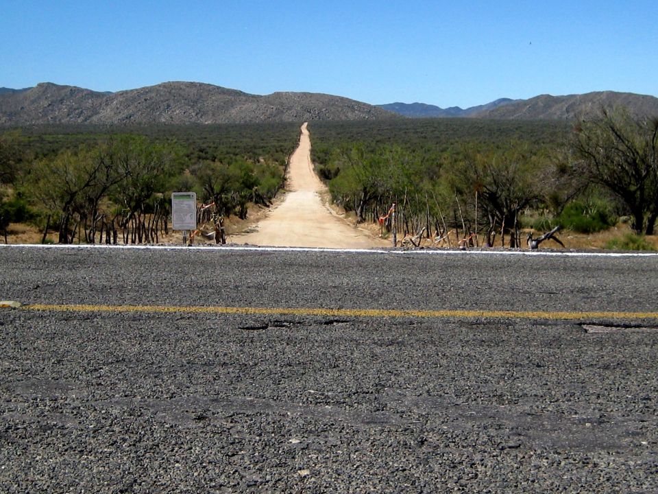 Gravel road leading to distant mountains under a clear blue sky, with trees and a signpost on the left
