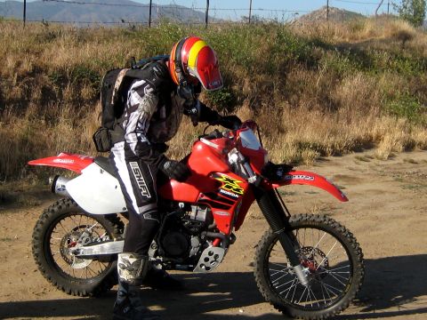 1996 Ford Bronco - A rider sits on a red Honda dirt bike on a dirt path with dry grass and hills in the background.