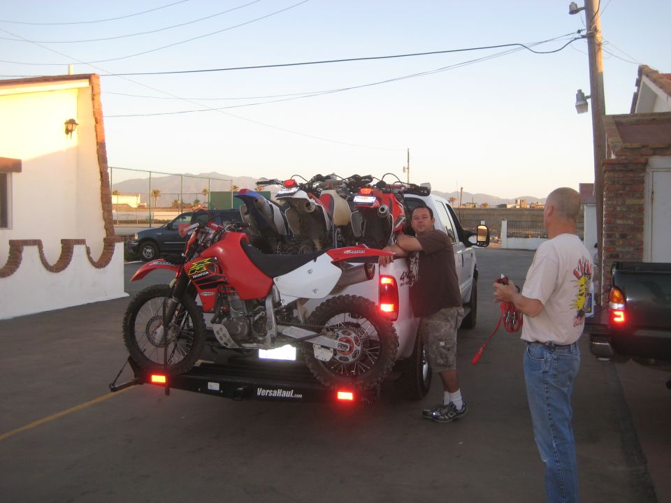 Pickup truck with a VersaHaul trailer carrying multiple dirt bikes. Two men stand nearby
