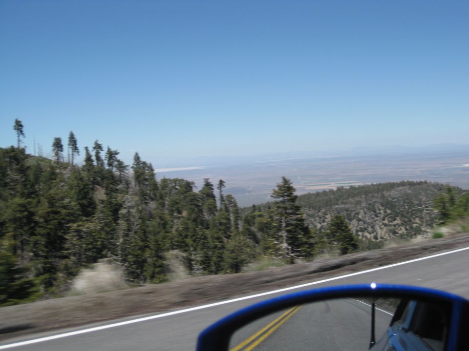 View from car window looking out at a mountain landscape with trees and a distant valley under a clear blue sky. A blue side mirror is visible in the foreground