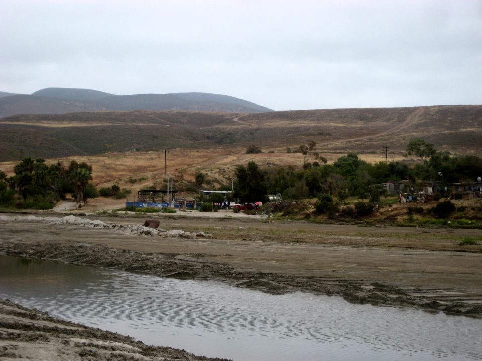 Overcast day shows a body of water in the foreground, with a dusty plain and some buildings in the midground. Hills are in the background