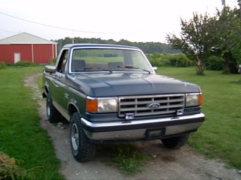 1987 Ford Bronco - This dark blue pickup truck, likely a 1987 model based on its grille, is parked on a dirt path in front of a red barn.