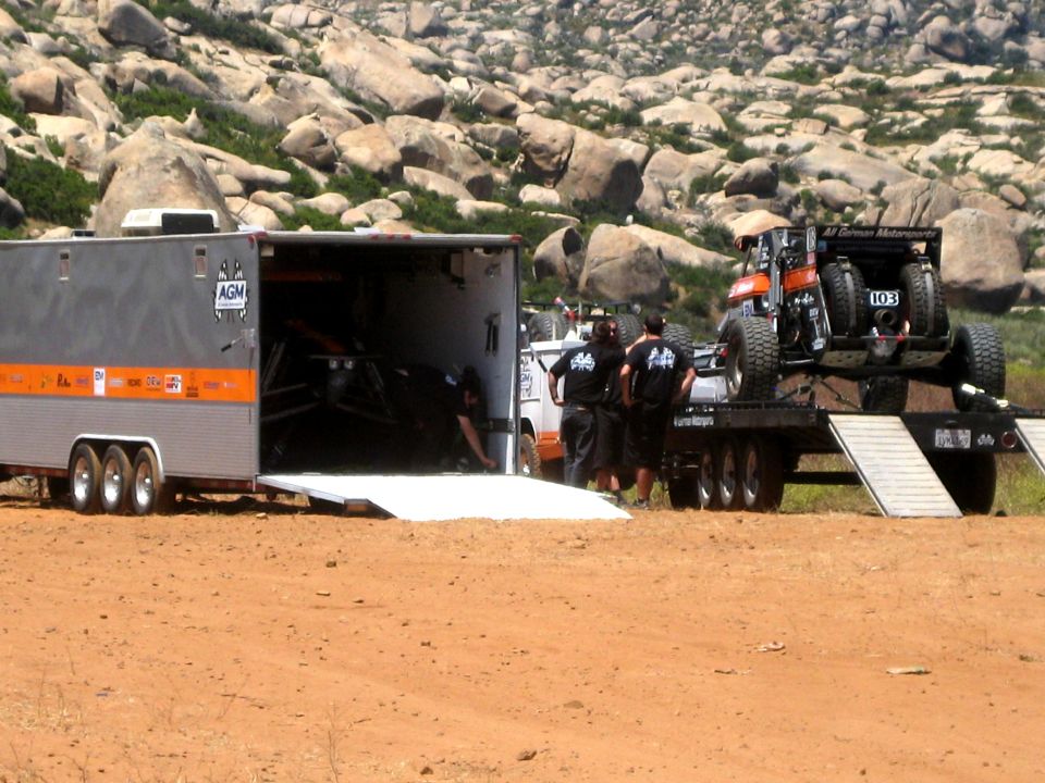 Off-road race buggy on a flatbed trailer, with a large enclosed trailer and people around