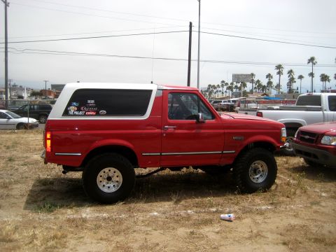 1996 Ford Bronco - A red Ford Bronco with a white top is parked on dry grass, with other vehicles and palm trees in the background.