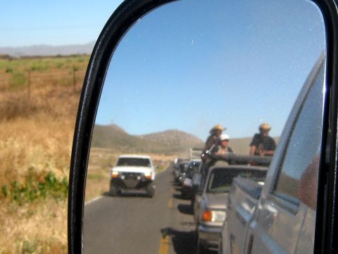 1996 Ford Bronco - A convoy of vehicles, including a white 1996 Ford Bronco, travels down a dusty road, as seen in a side mirror.