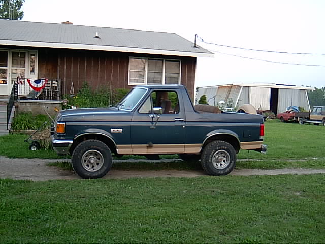 Dark green two-tone Ford Bronco with top partially removed, lifted on off-road tires, parked by a rural house